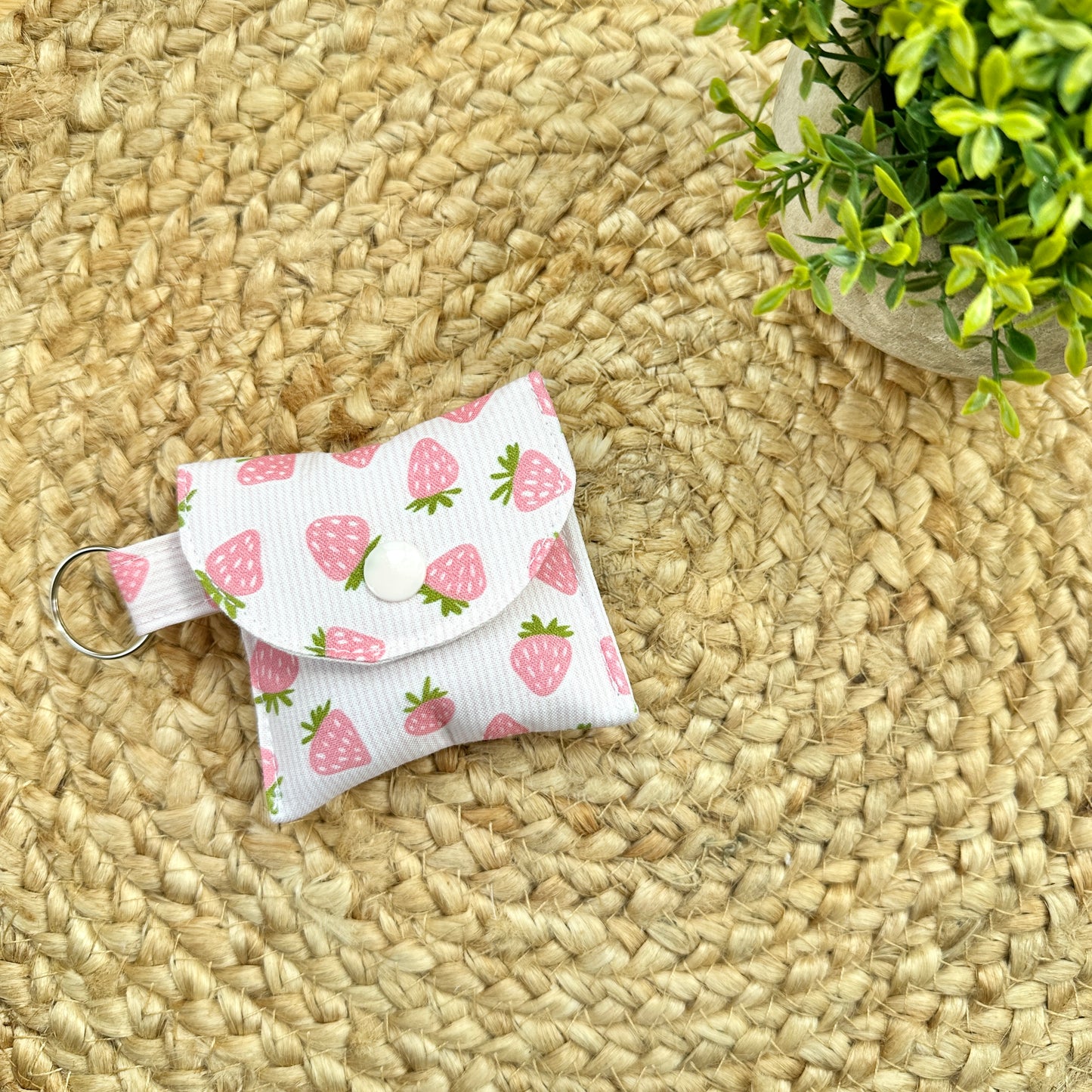 A small fabric pouch with a pink strawberry print on a white background, featuring a snap-button closure and a metal key ring, placed on a woven straw mat with a small potted plant in the corner.