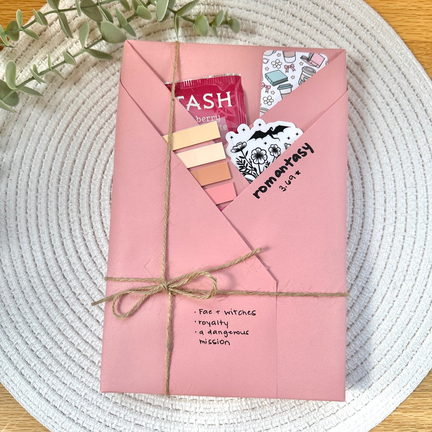 a pink wrapped romantasy book on a woven mat with decorative stationery items
