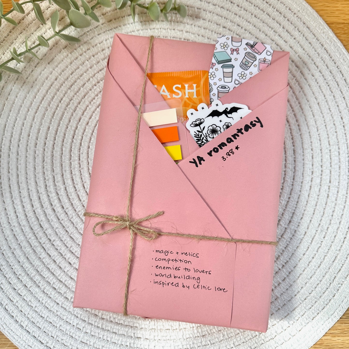 a pink wrapped YA romantasy book on a woven mat with decorative stationery items