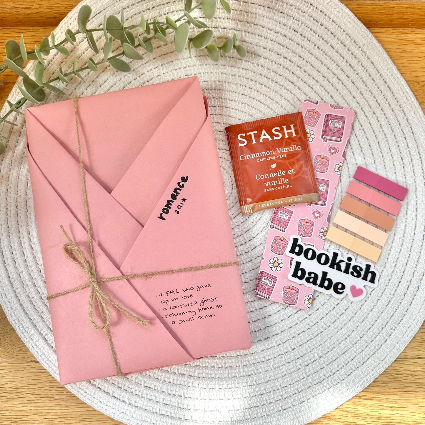 a pink wrapped romance book on a woven mat with tea bag, bookmark, annotative sticker tabs, and bookish babe sticker