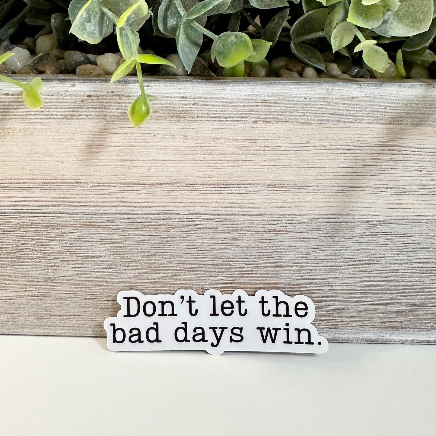 A close-up of a mental health decal that reads 'Don't let the bad days win.' on a white surface with a plant in the background.