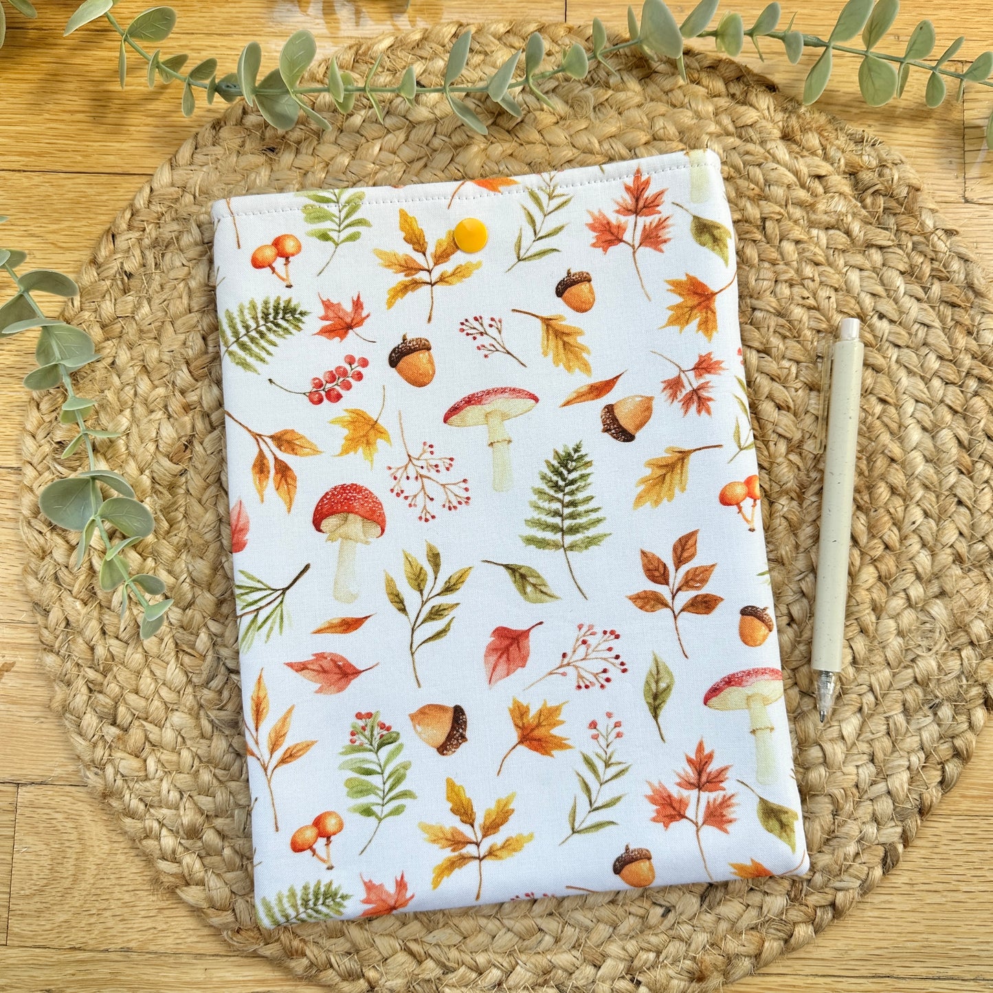 A fabric book sleeve with an autumn-themed print of mushrooms, acorns, and leaves on a white background, styled on a woven placemat with a pen and eucalyptus leaves.