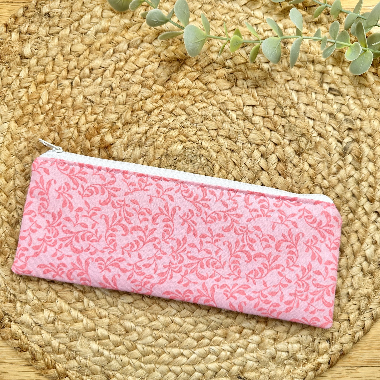 A zippered cotton pencil pouch with a pink floral damask pattern, resting on a round woven straw mat with a green eucalyptus sprig in the background.