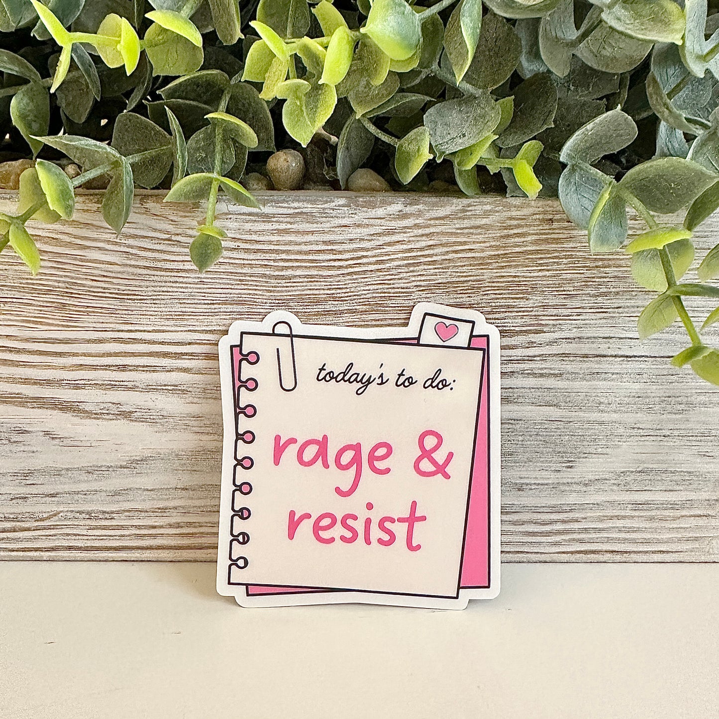 Notebook-style sticker with 'today's to do: rage & resist' text on a wooden surface.