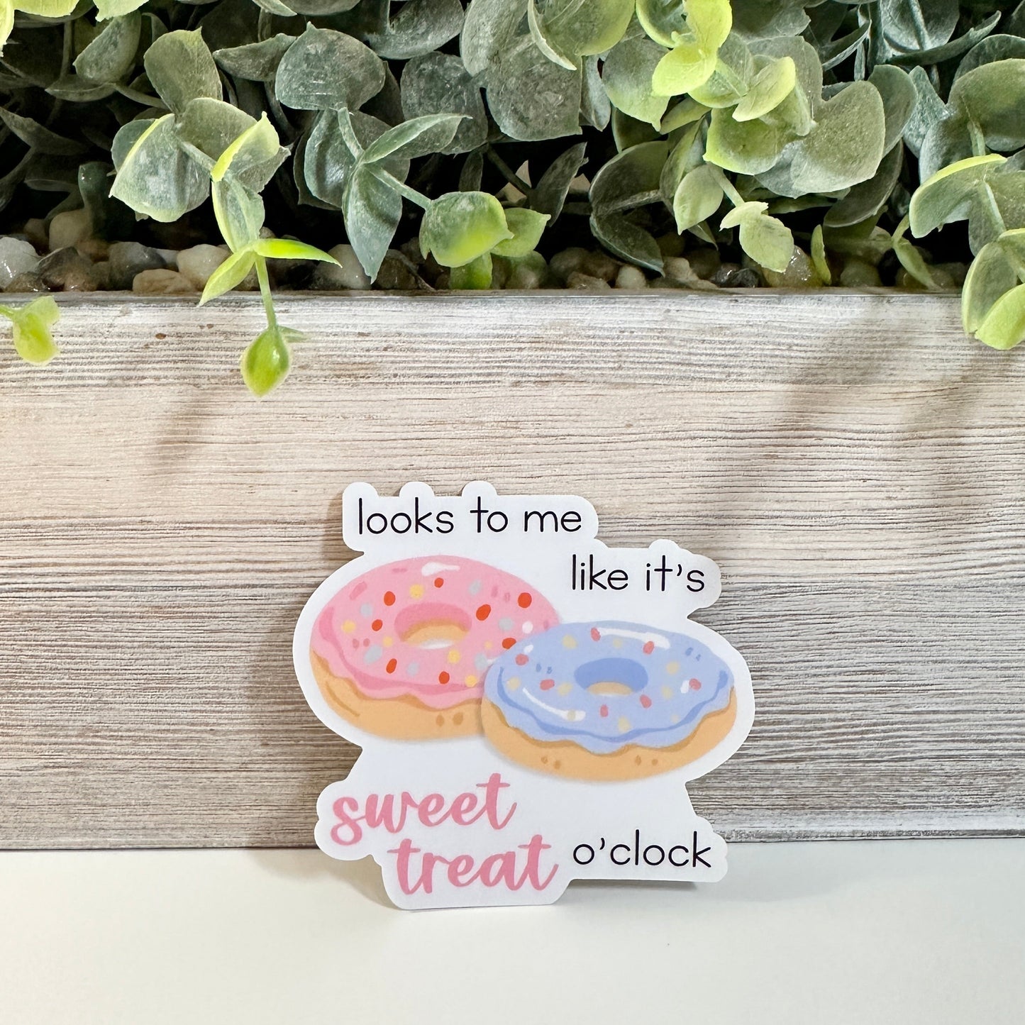 A waterproof matte vinyl decal featuring two donuts with the text 'looks to me like it's sweet treat o'clock', displayed on a light surface in front of a wooden planter with green leaves.