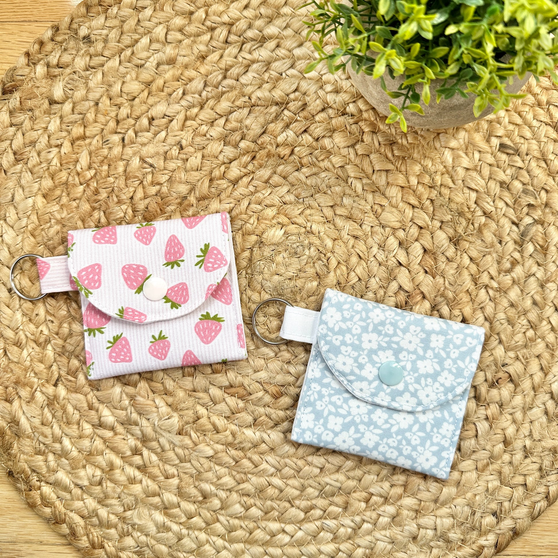 Two small fabric card holders or coin pouches with key rings, displayed on a round woven straw mat. One pouch has a white background with a pink strawberry pattern, and the other has a light blue background with a white floral pattern.