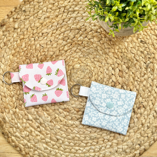 Two small fabric card holders or coin pouches with key rings, displayed on a round woven straw mat. One pouch has a white background with a pink strawberry pattern, and the other has a light blue background with a white floral pattern.