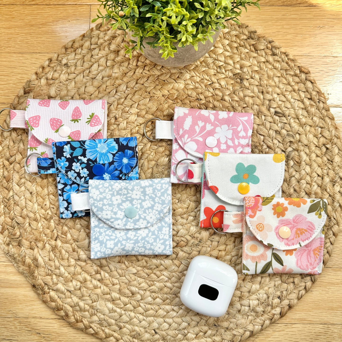 Six small fabric card holders or pouches with floral and fruit prints are arranged on a round woven placemat over a wood surface, with a small potted plant above and a white earbud case below.