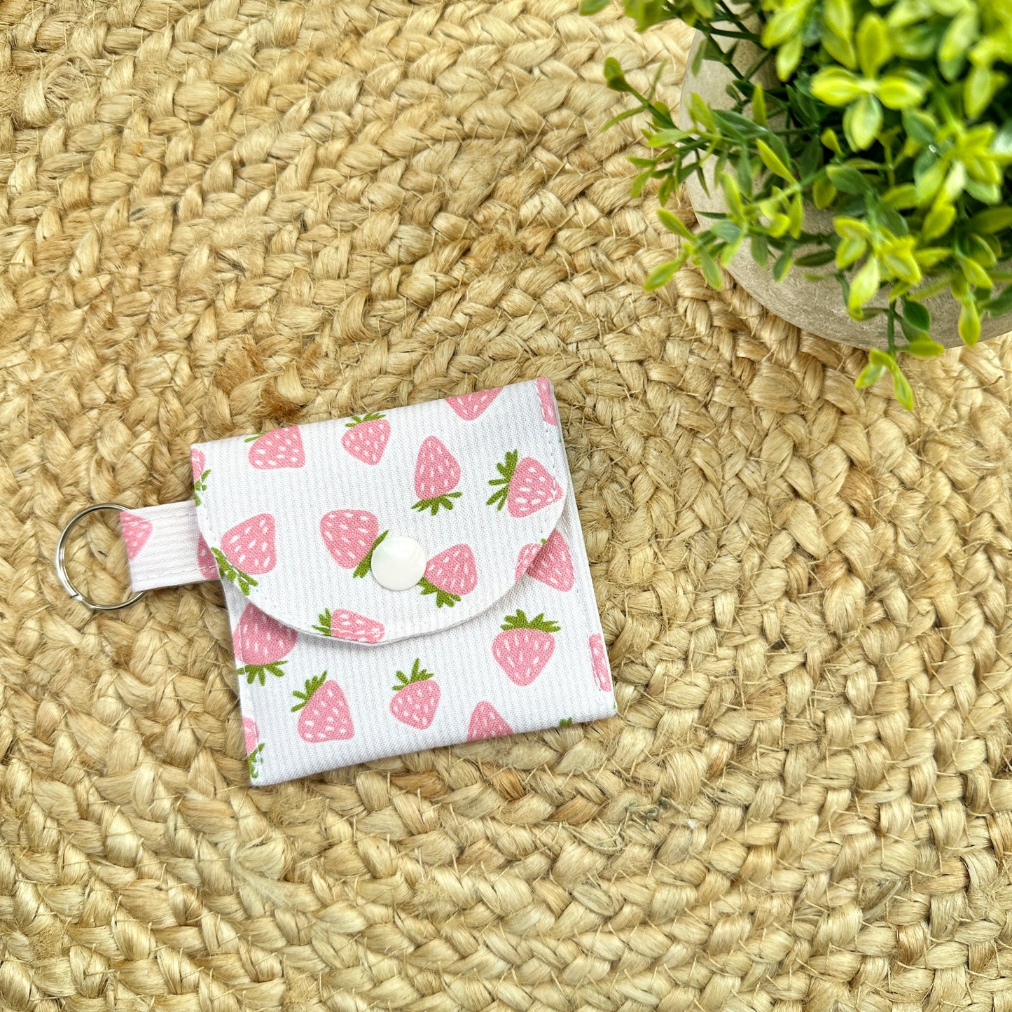 A small fabric pouch with a pink strawberry print on a white striped background, featuring a snap button closure and a metal key ring, placed on a woven straw mat.