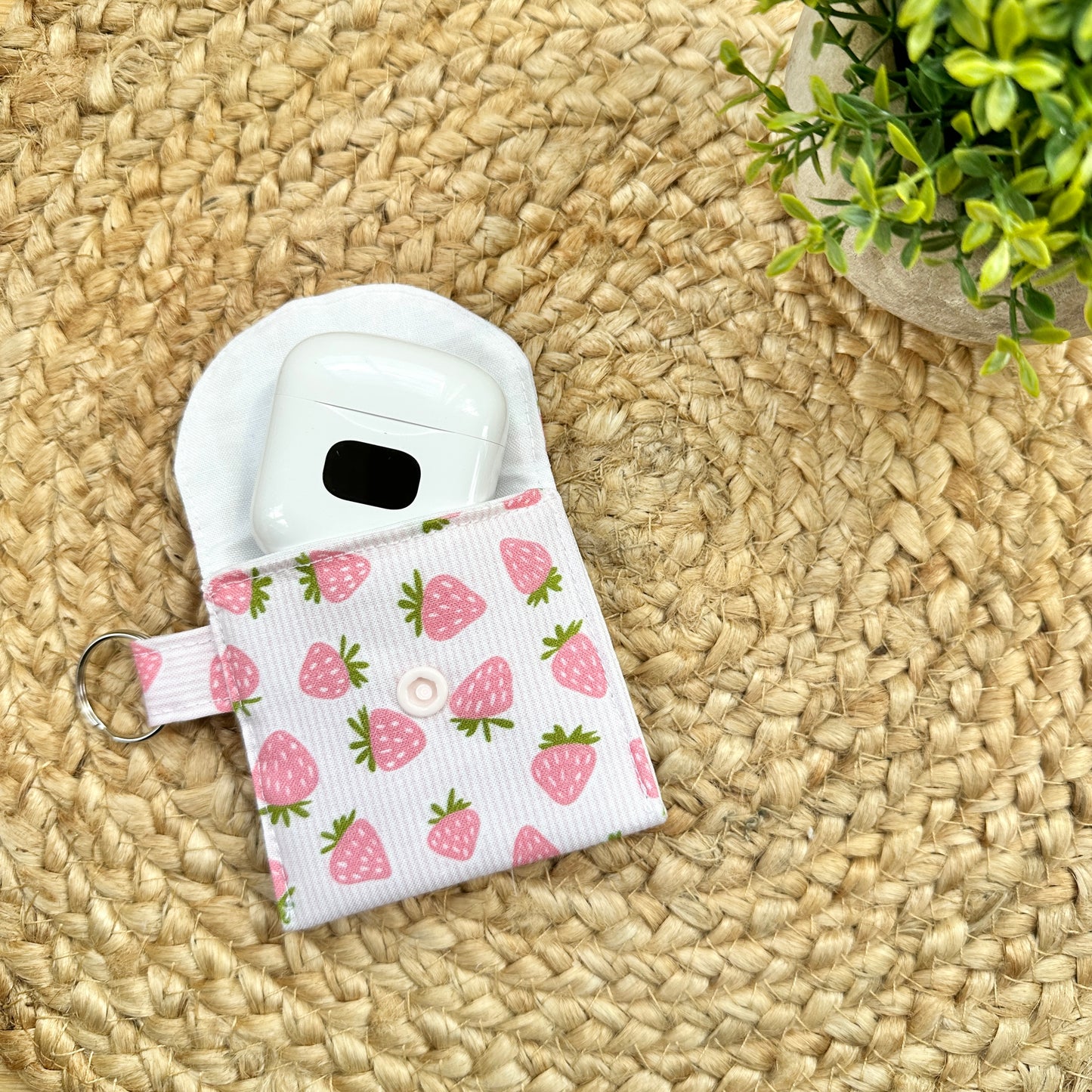 A fabric AirPods case with a pink strawberry print and a white snap button, shown holding a white AirPods case on a woven straw mat with a small green plant in the corner.