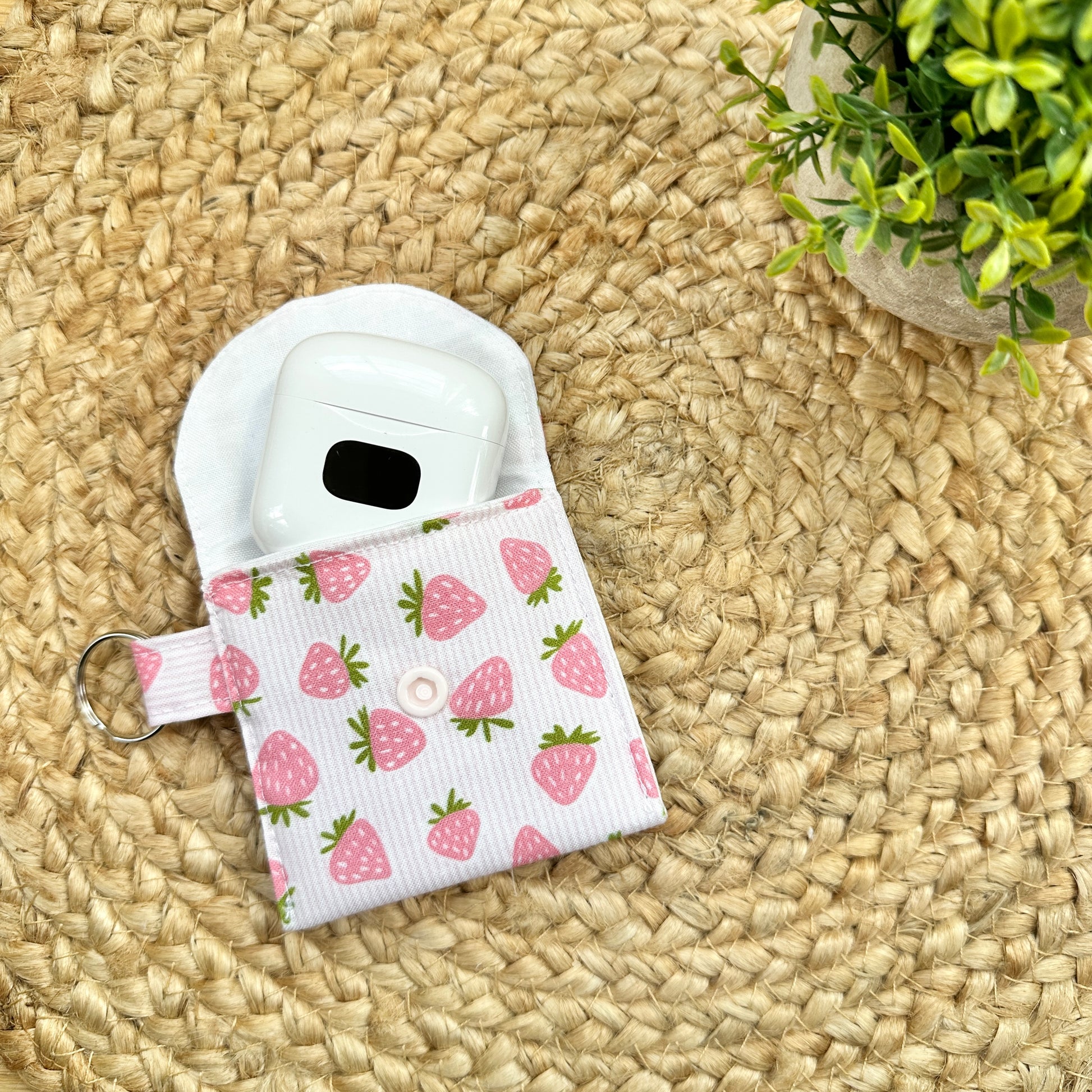A fabric AirPods case with a pink strawberry print and a white snap button, shown holding a white AirPods case on a woven straw mat with a small green plant in the corner.