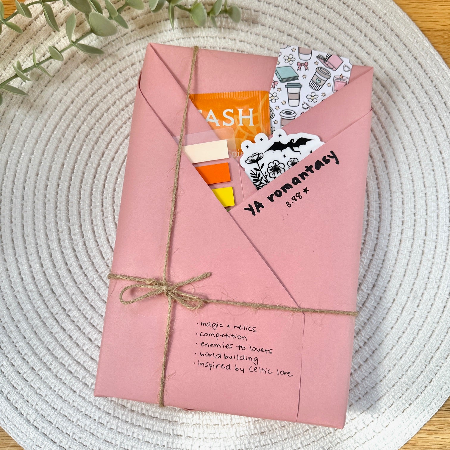 a pink wrapped YA romantasy book on a woven mat with decorative stationery items