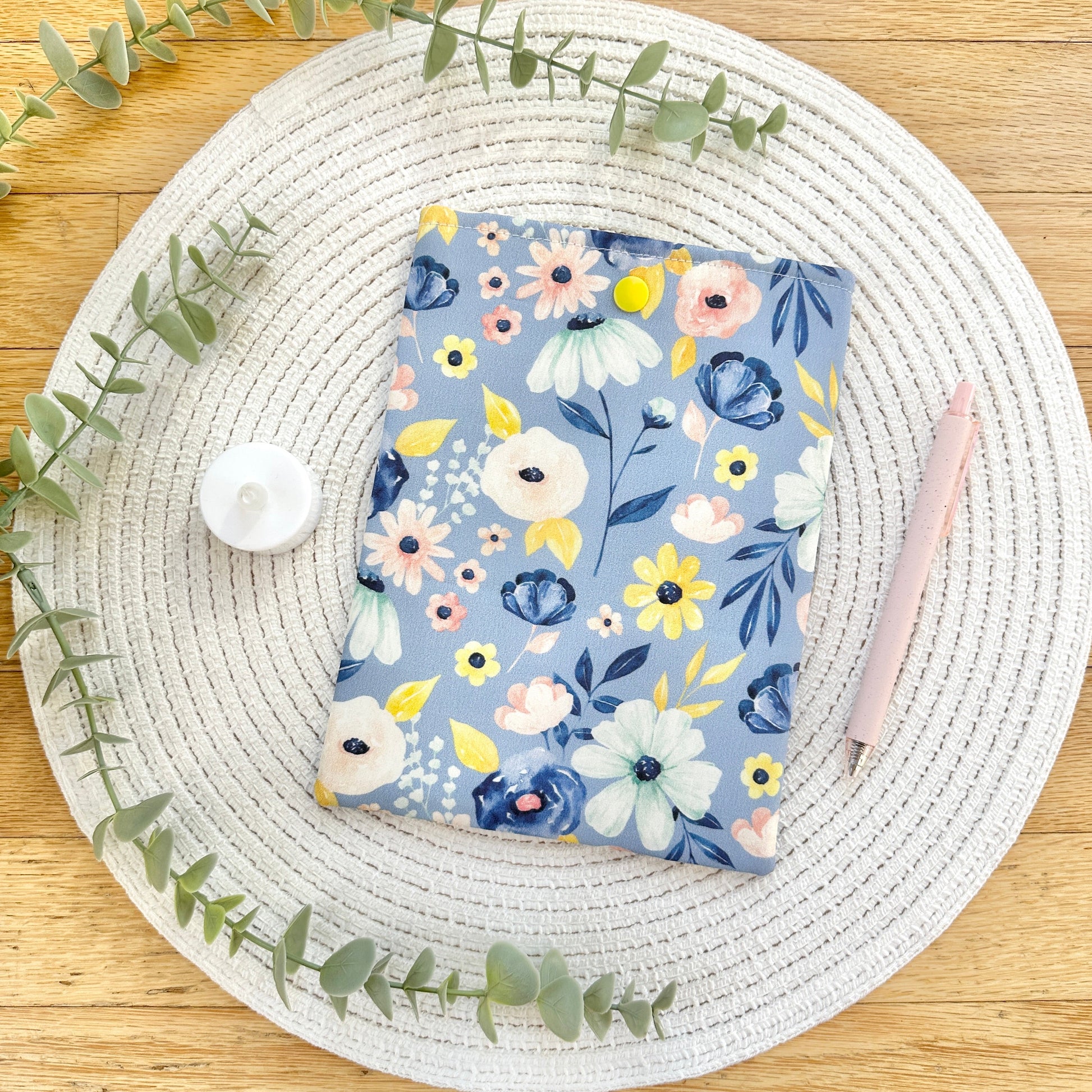 Floral-patterned kindle sleeve on a woven placemat with eucalyptus leaves.
