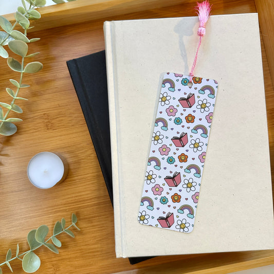 Bookmark with colorful design of books, rainbows, and flowers on a book on a wooden surface with a candle and plant.