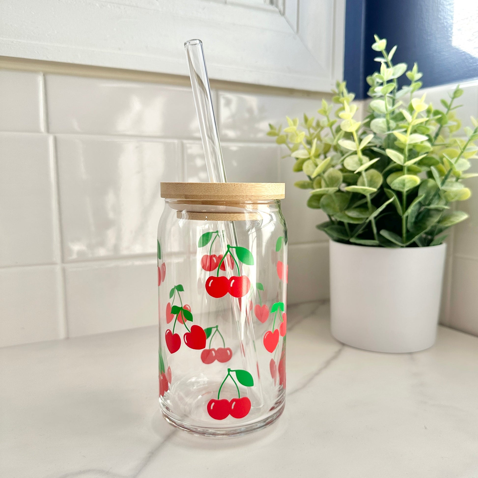 Clear glass coffee cup with cherry design, wooden lid, and straw on a kitchen counter.