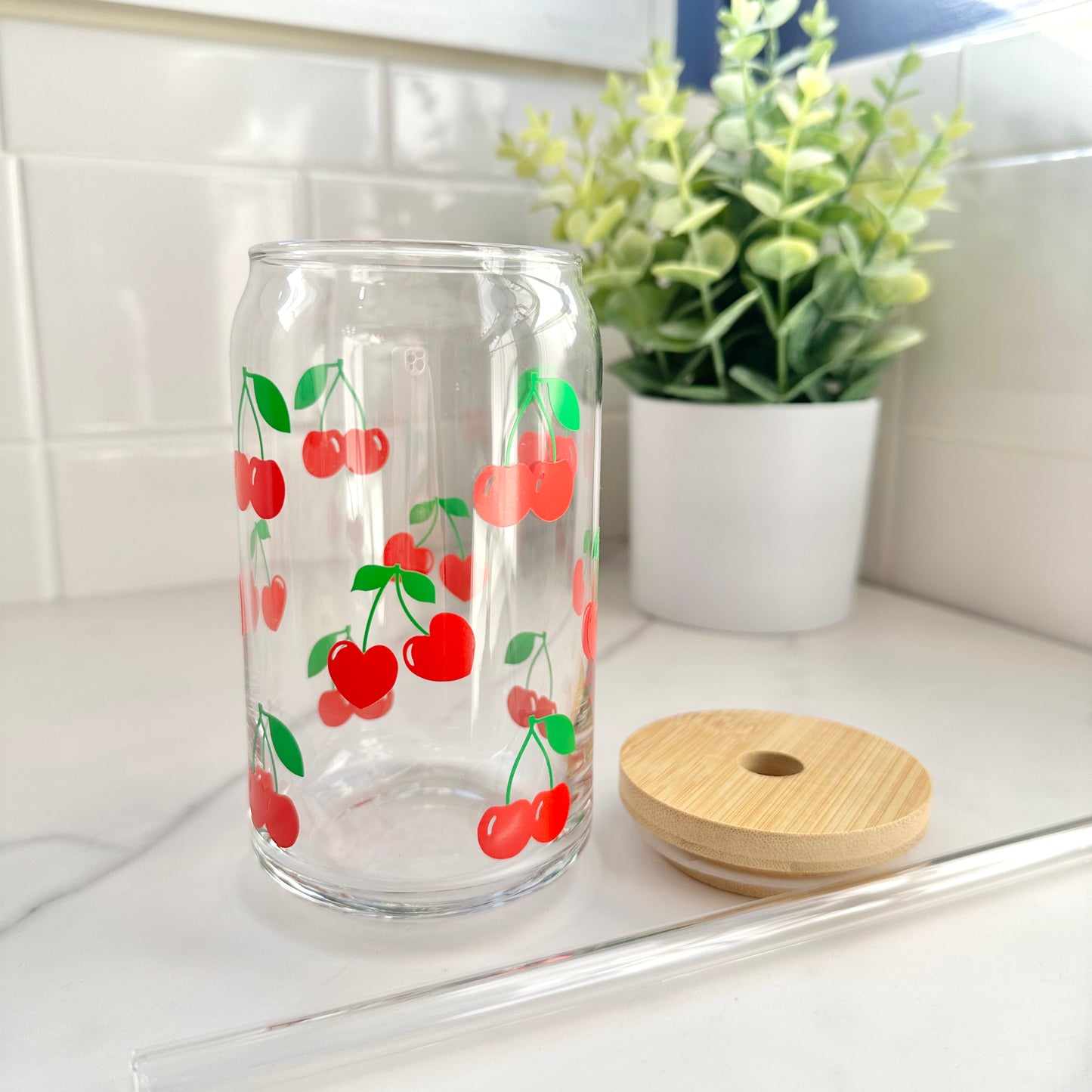 cherry glass coffee cup with wood top and glass straw on counter with plant in the background