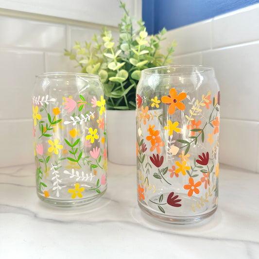 Two clear glass can-shaped tumblers decorated with colorful floral and leaf patterns, displayed on a white marble countertop in front of a white tiled backsplash with a green plant in the background.