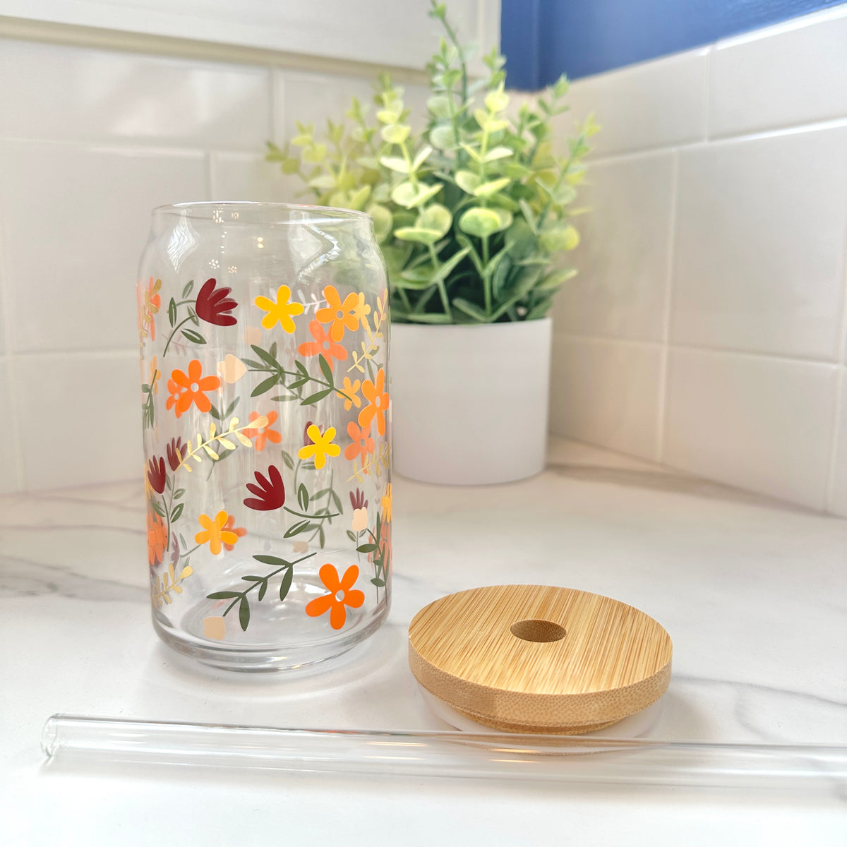 A clear glass can-shaped tumbler with a colorful fall floral pattern, shown on a white countertop with its bamboo lid and clear glass straw in front, and a potted plant in the background.