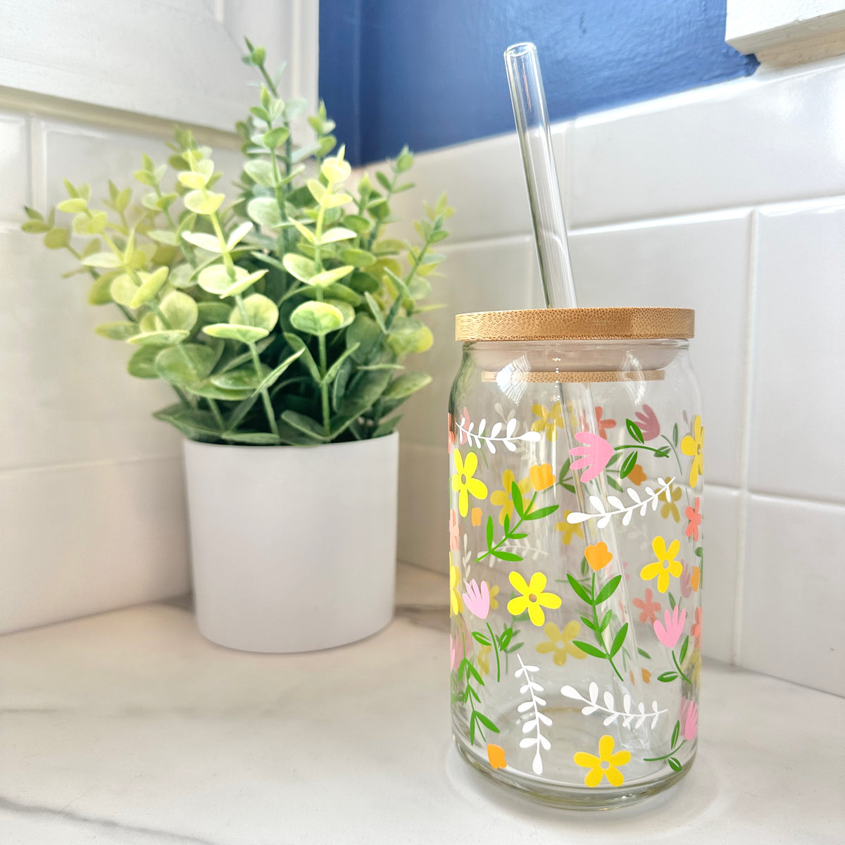 A floral-patterned iced coffee glass up with a bamboo lid and a clear glass straw, set on a white tiled countertop next to a potted green plant.