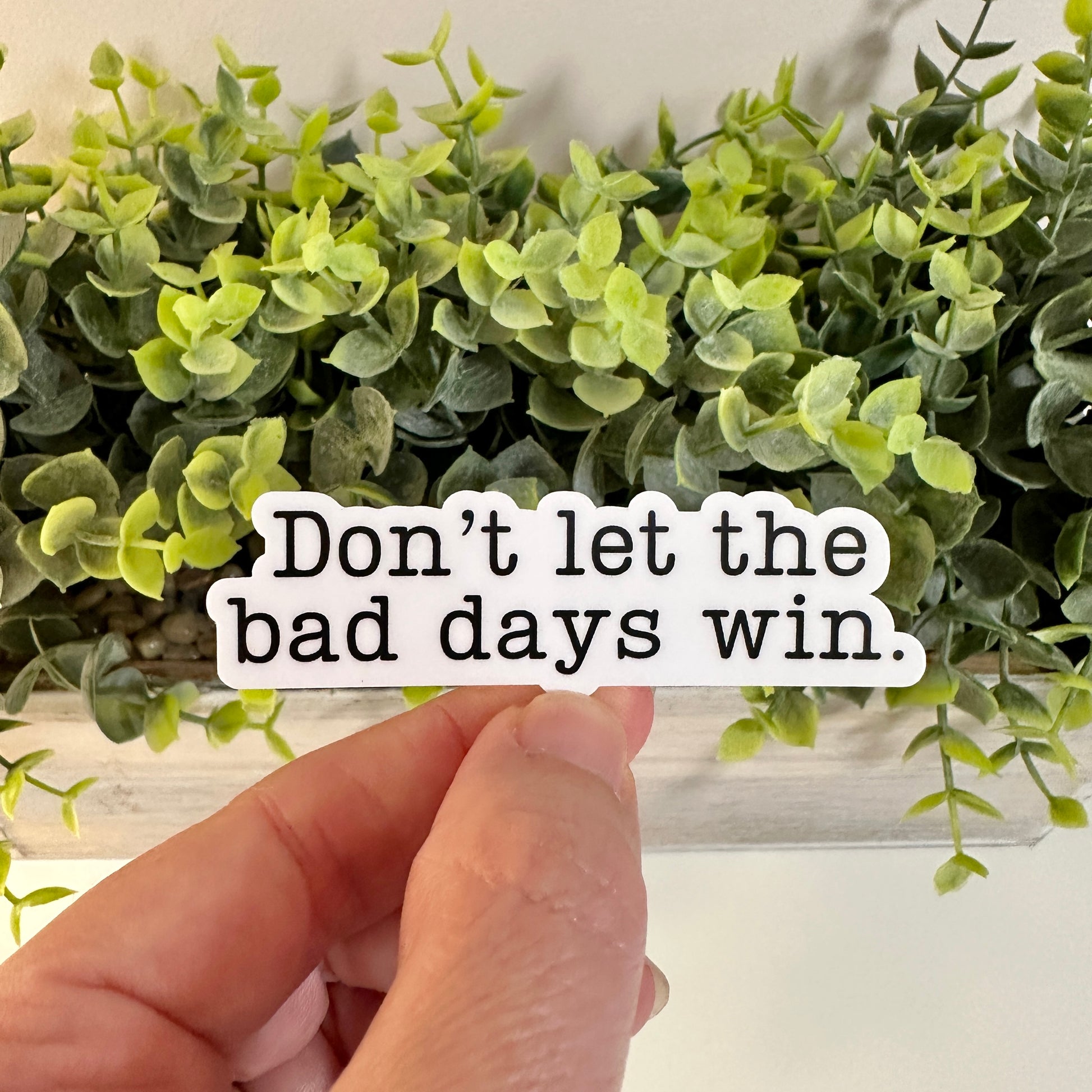 A hand holds a white die-cut sticker with the text 'Don’t let the bad days win.' in black serif lettering, positioned in front of a green artificial plant arrangement.
