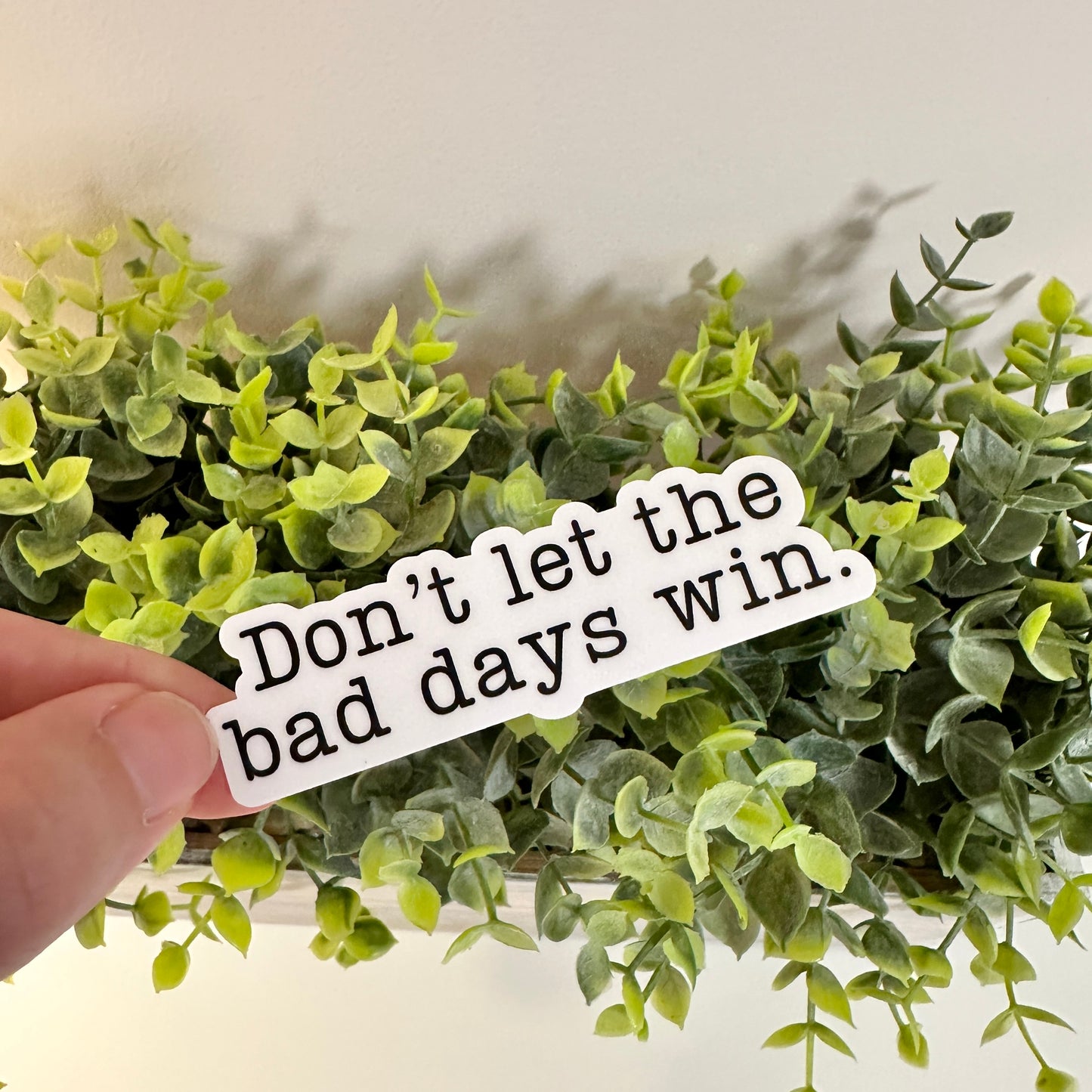 A hand holds a waterproof mental health sticker with the text 'Don't let the bad days win.' in front of a green plant.