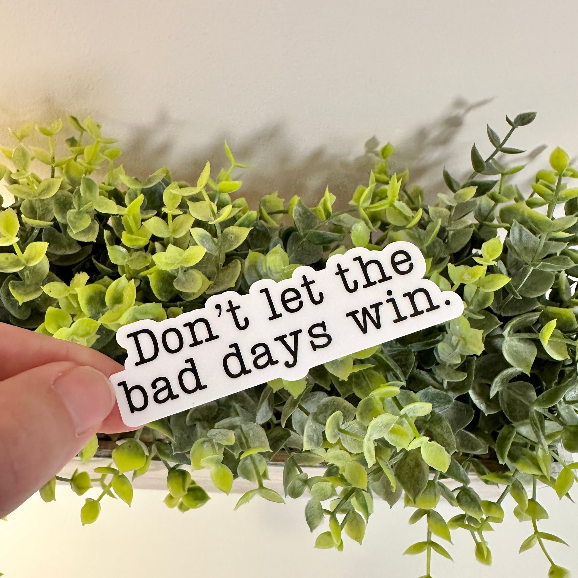 A hand holds a waterproof mental health sticker with the text 'Don't let the bad days win.' in front of a green plant.