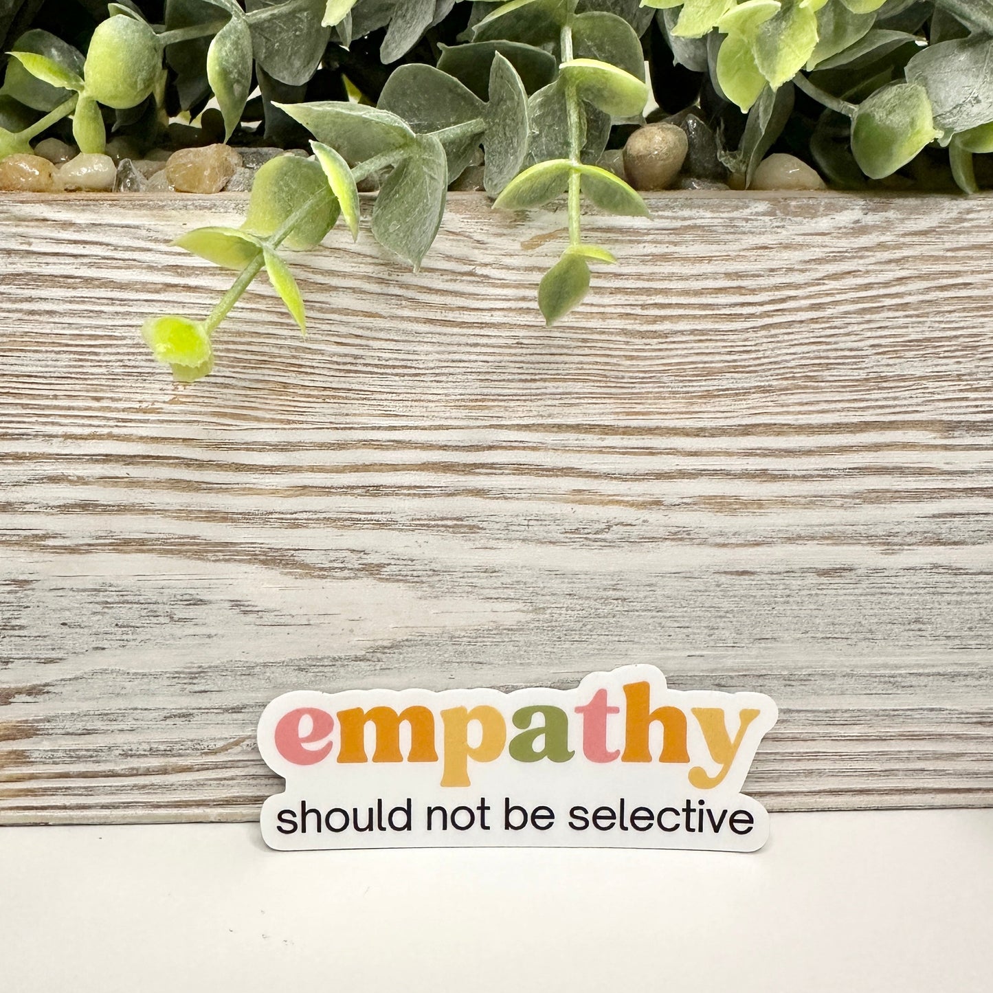 Sticker with 'empathy should not be selective' text on a wooden surface with greenery.