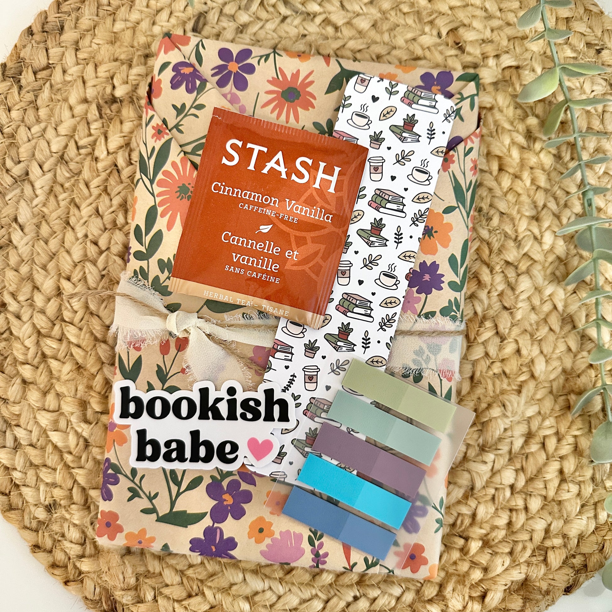 A styled flat-lay of the items included with the cozy blind dte with a book featuring a floral-patterned book wrap, a Stash cinnamon vanilla coffee bag, a coffee-themed bookmark, and a set of colorful book tabs, all arranged on a woven straw mat.