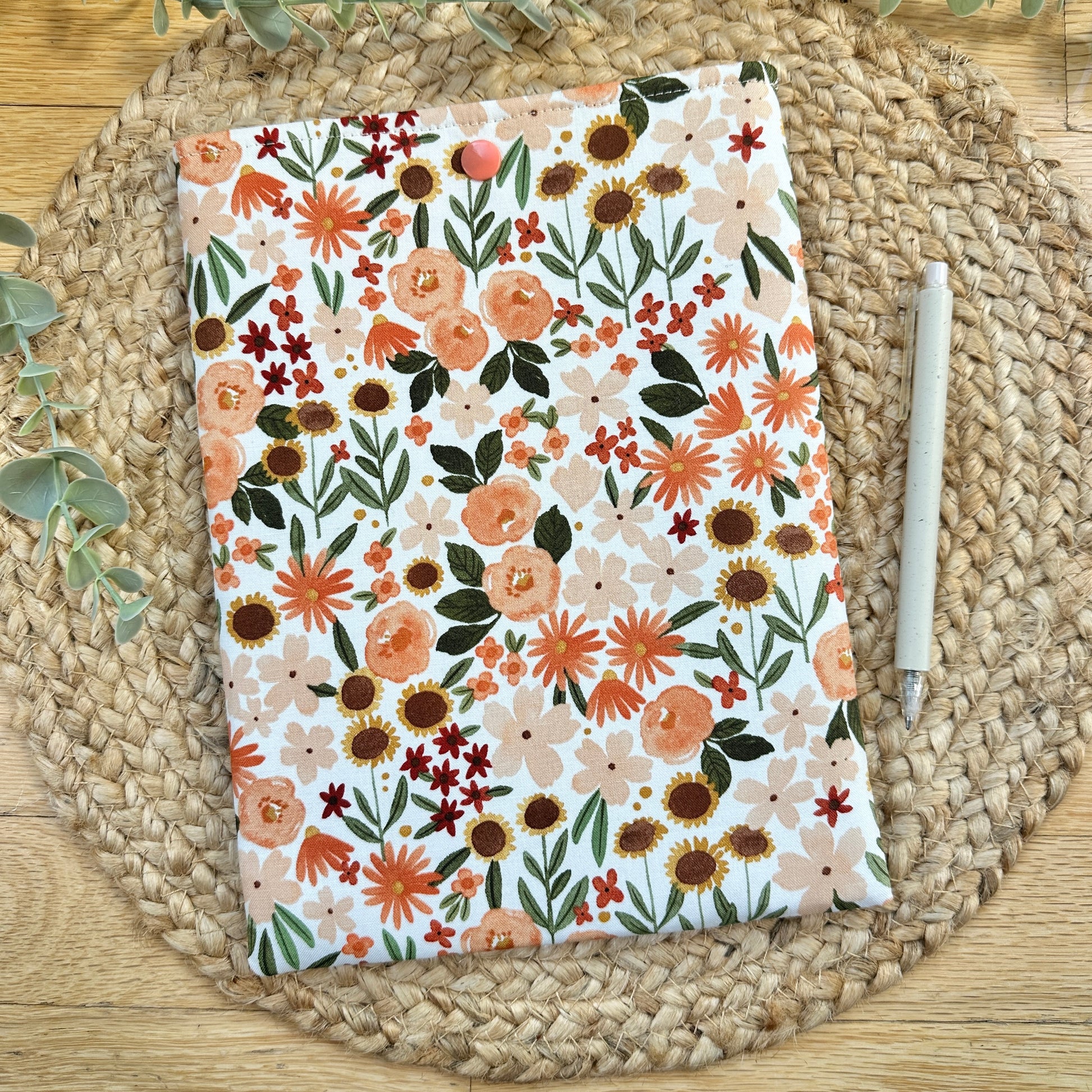 A fabric paperbook sleeve with a colorful fall floral print, displayed on a round woven placemat with a white pen beside it.