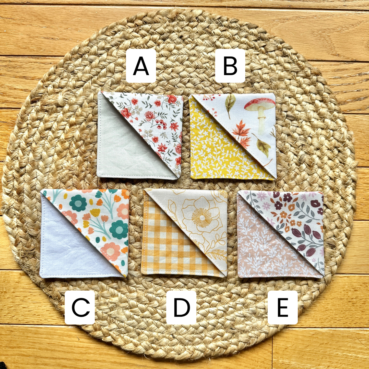 Five fall-themed fabric corner bookmarks by Alyssa Mae Crafts are arranged on a round woven placemat over a wood floor. Each bookmarkj is labeled with a white letter sticker (A, B, C, D, E).