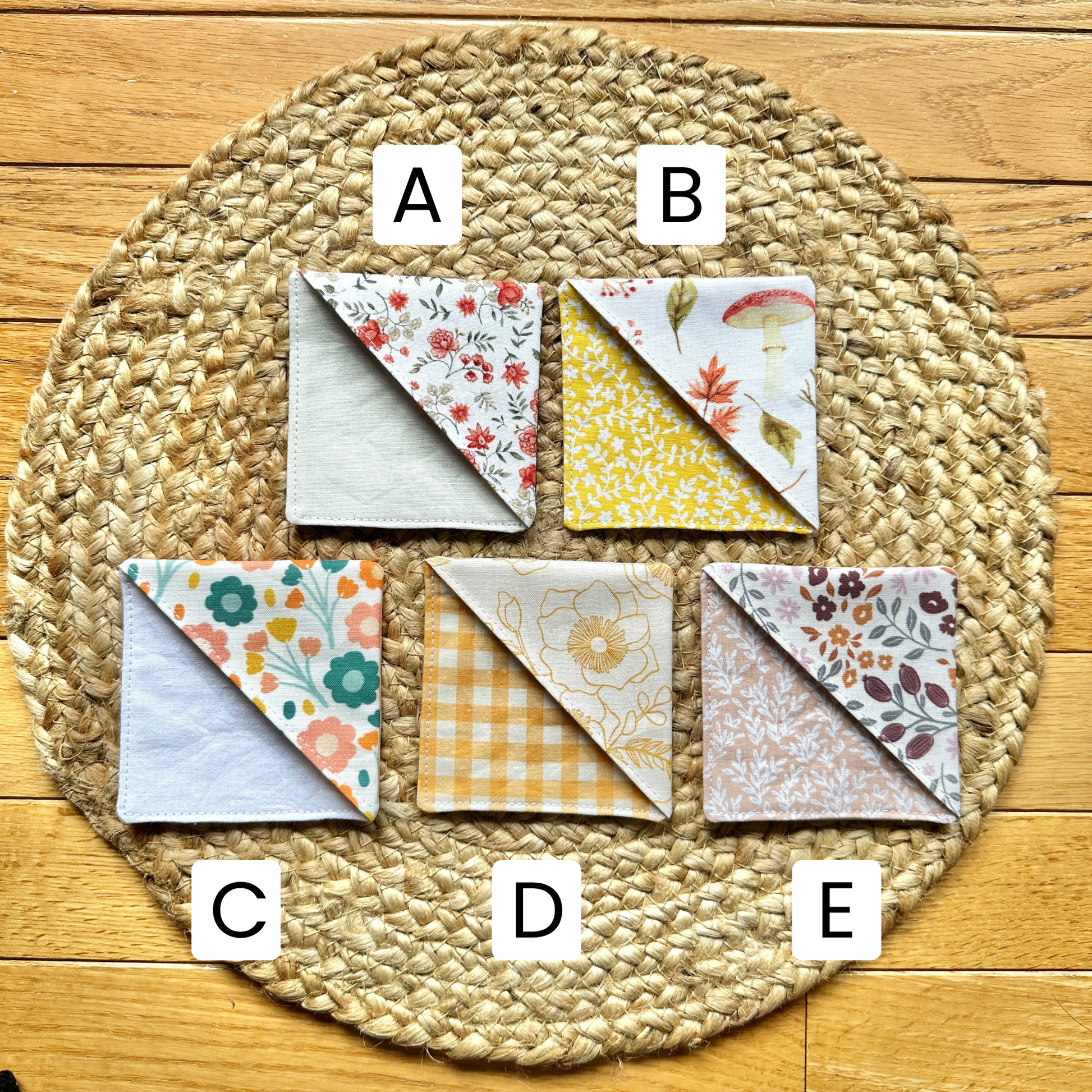 Five fall-themed fabric corner bookmarks by Alyssa Mae Crafts are arranged on a round woven placemat over a wood floor. Each bookmarkj is labeled with a white letter sticker (A, B, C, D, E).