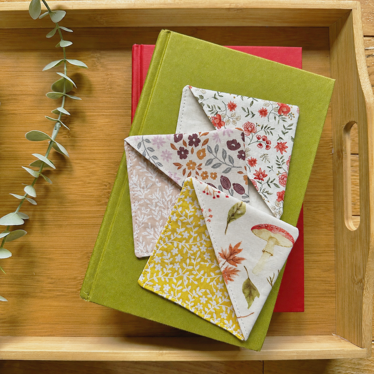 Three fabric corner bookmarks with floral fall prints are arranged on top of a green book, which sits on a red book inside a light wood tray. A sprig of eucalyptus is visible along the left side.