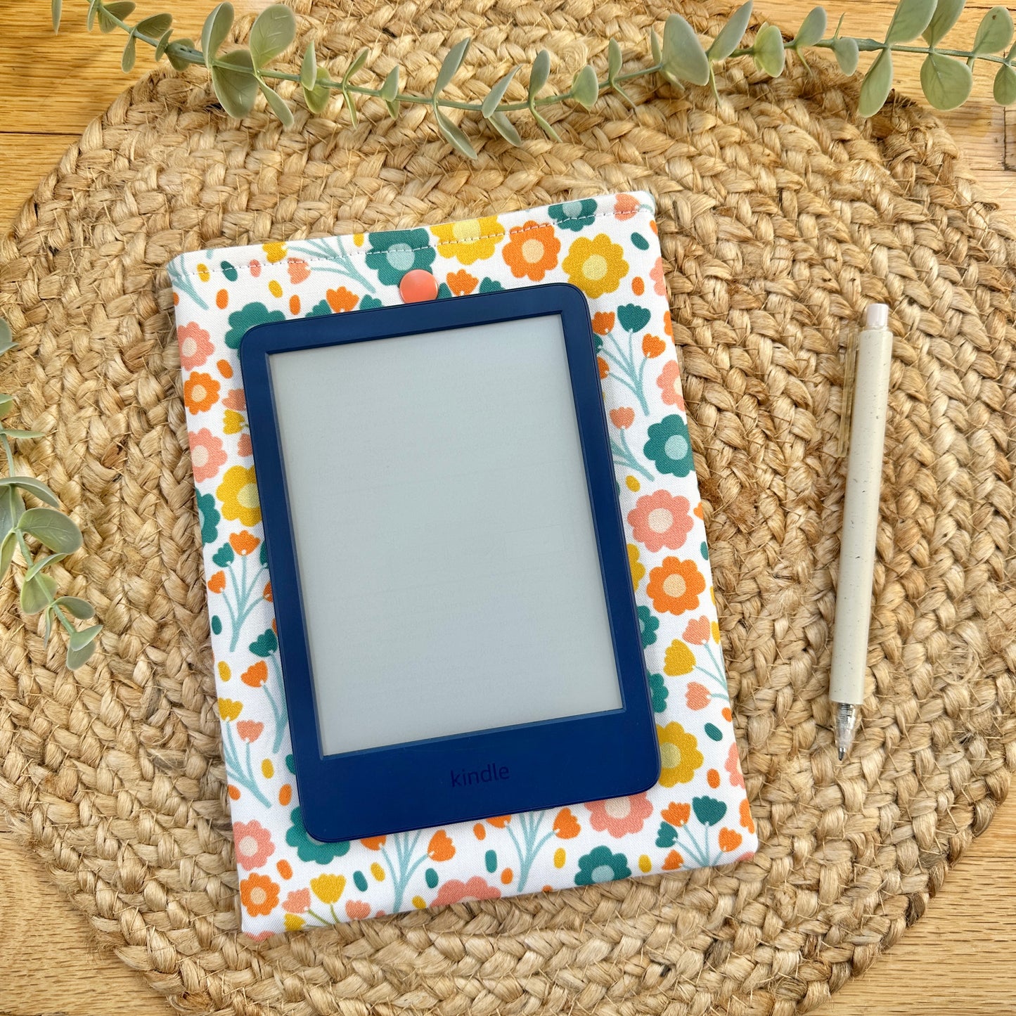 A fabric tablet sleeve with a colorful retro autumnal floral pattern, holding an e-reader, styled on a round woven straw placemat over a wooden table with eucalyptus sprigs.