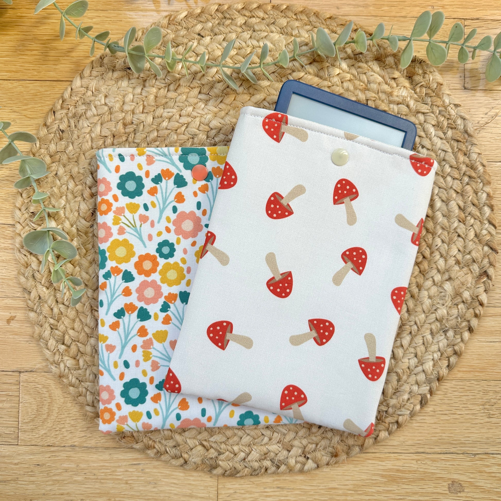 Two fabric tablet sleeves displayed on a woven round mat on a wooden surface. The front sleeve is white with a red mushroom print, and the sleeve behind it has a multicolor floral pattern.