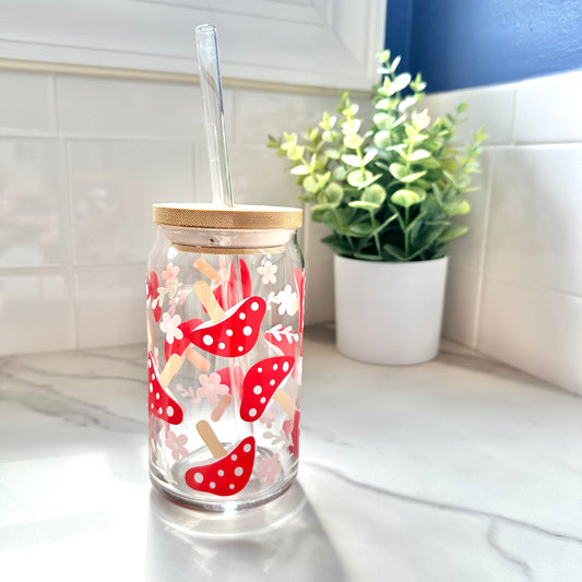 A clear glass tumbler with a bamboo lid and clear straw, decorated with red and white mushrooms and pink flowers, sitting on a marble countertop with a potted plant in the background.