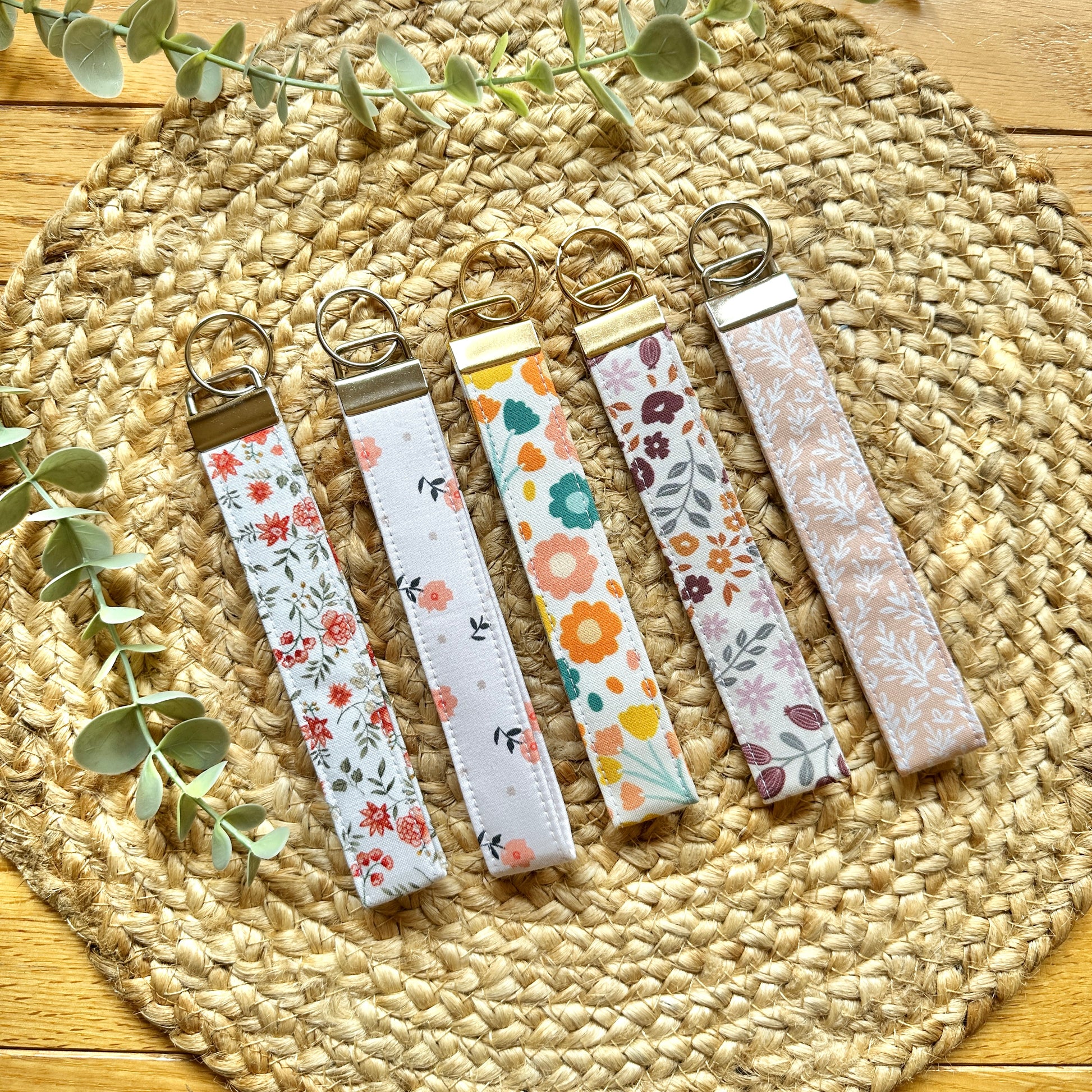 Five fabric wristlet keychains with metal key rings are arranged on a round woven straw mat. The keychains have different autumn floral and botanical prints in soft colors including pink, orange, green, blue, and purple on white and beige backgrounds.