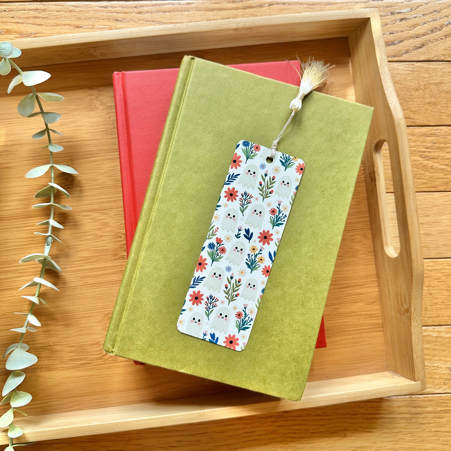 A rectangular paper bookmark with a colorful floral and ghost pattern and a tassel, resting on top of a green book which is stacked on a red book inside a bamboo tray.