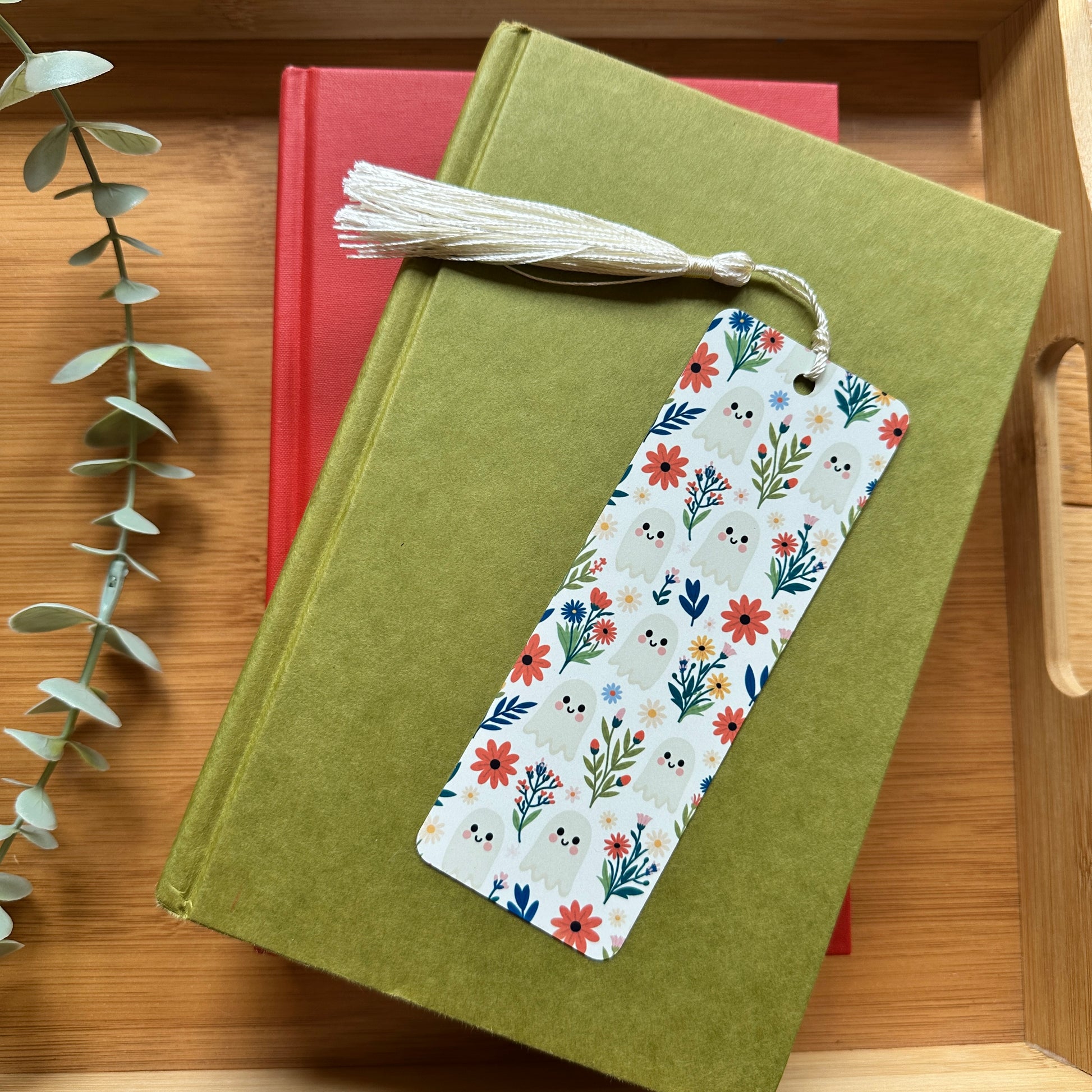 A rectangular bookmark with a white background and a colorful floral and ghost print, featuring a cream tassel, resting on top of an green book on a wooden surface.