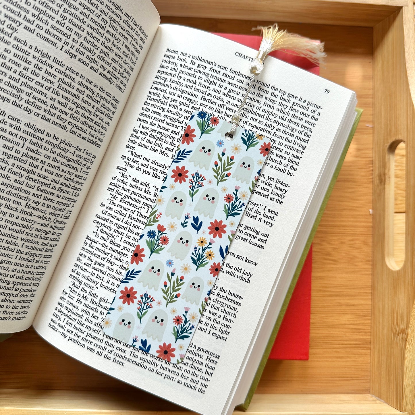 A decorative fall/Halloween metal bookmark with tassel, shown resting on an open book inside a wooden tray.