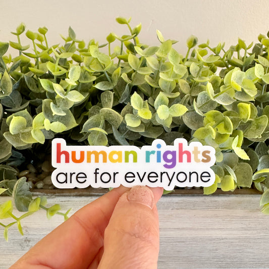 Hand holding a 'human rights are for everyone' sticker with green leaves in the background