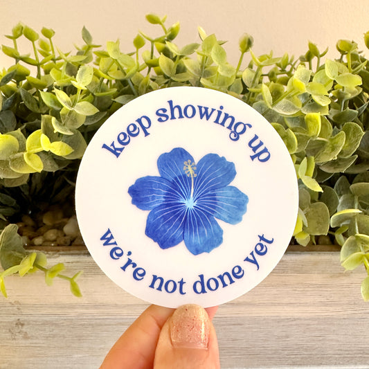 Circular sticker with blue flower and text 'keep showing up, we're not done yet' held in front of green foliage.