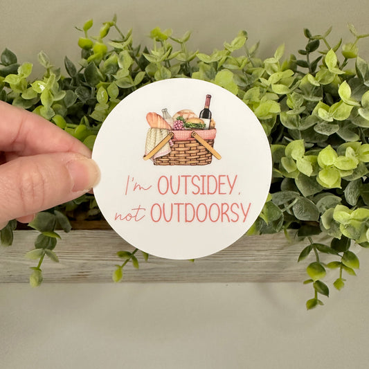 hand holding sticker with text "I'm outsidey, not outdoorsy" and picture of picnic basket