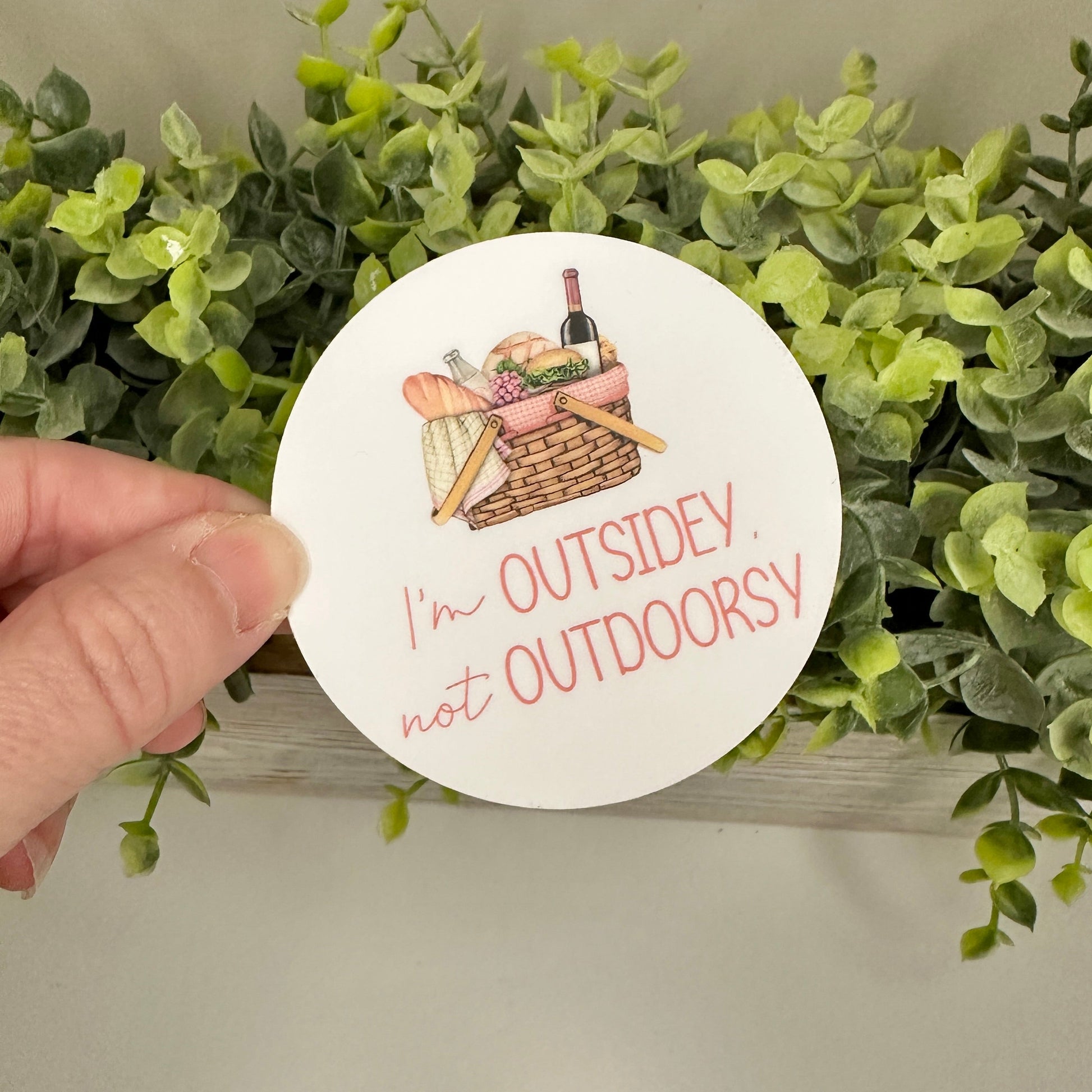 Circular sticker with picnic illustration and text 'I'm Outsidey, not Outdoorsy' held over green foliage.