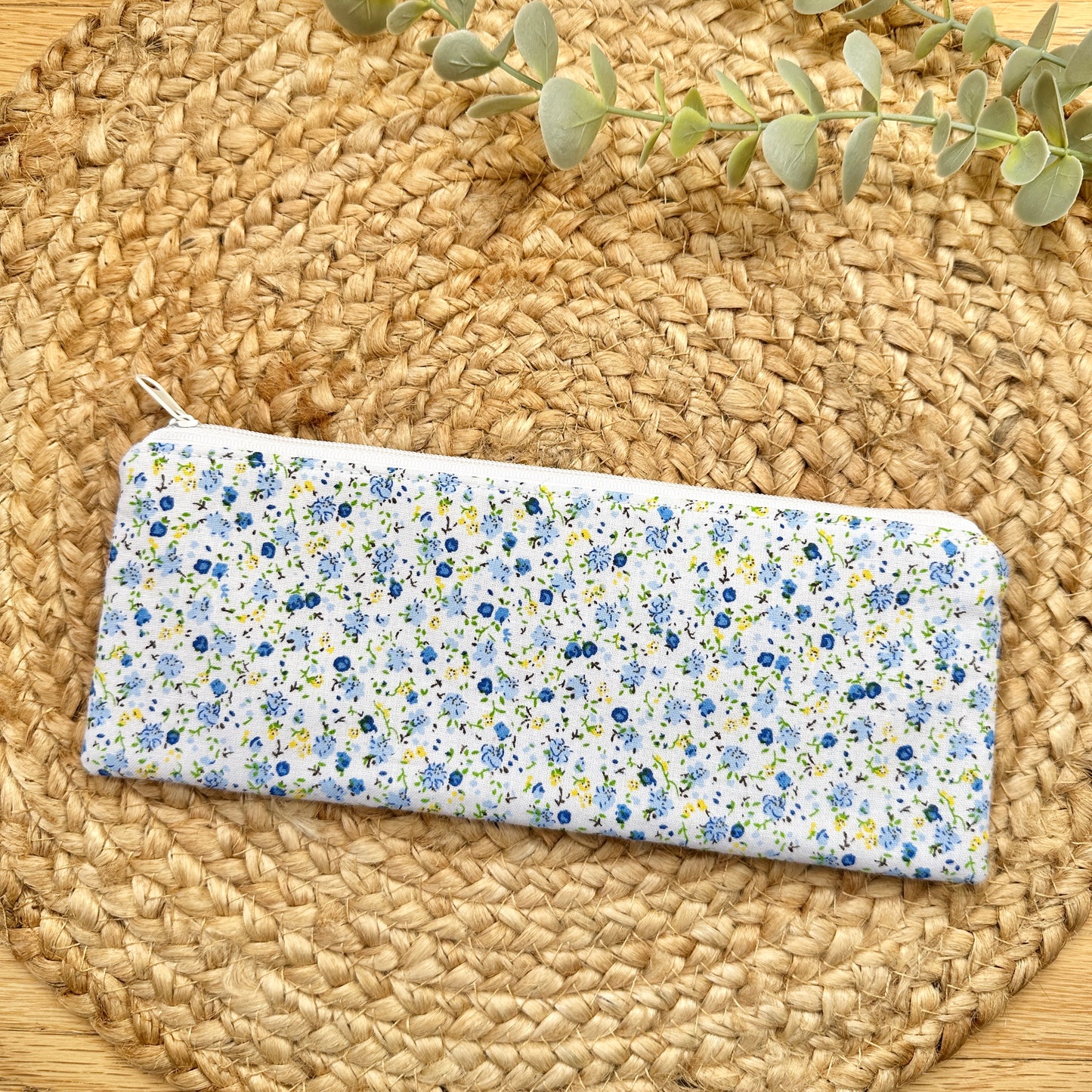 A rectangular zip pouch with a white background and a dense blue, yellow, and green floral print, placed on a round woven straw mat with a sprig of eucalyptus at the top.