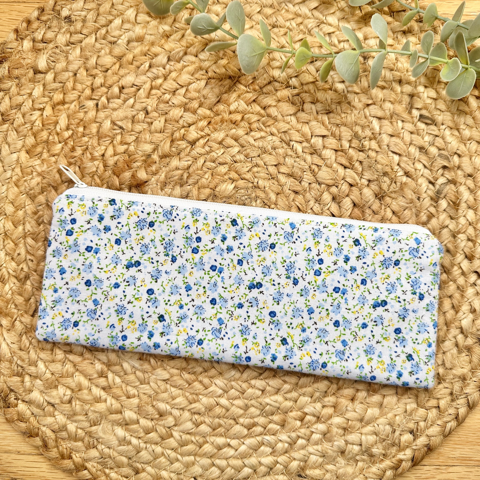A rectangular zip pouch with a white background and a dense blue, yellow, and green floral print, placed on a round woven straw mat with a sprig of eucalyptus at the top.