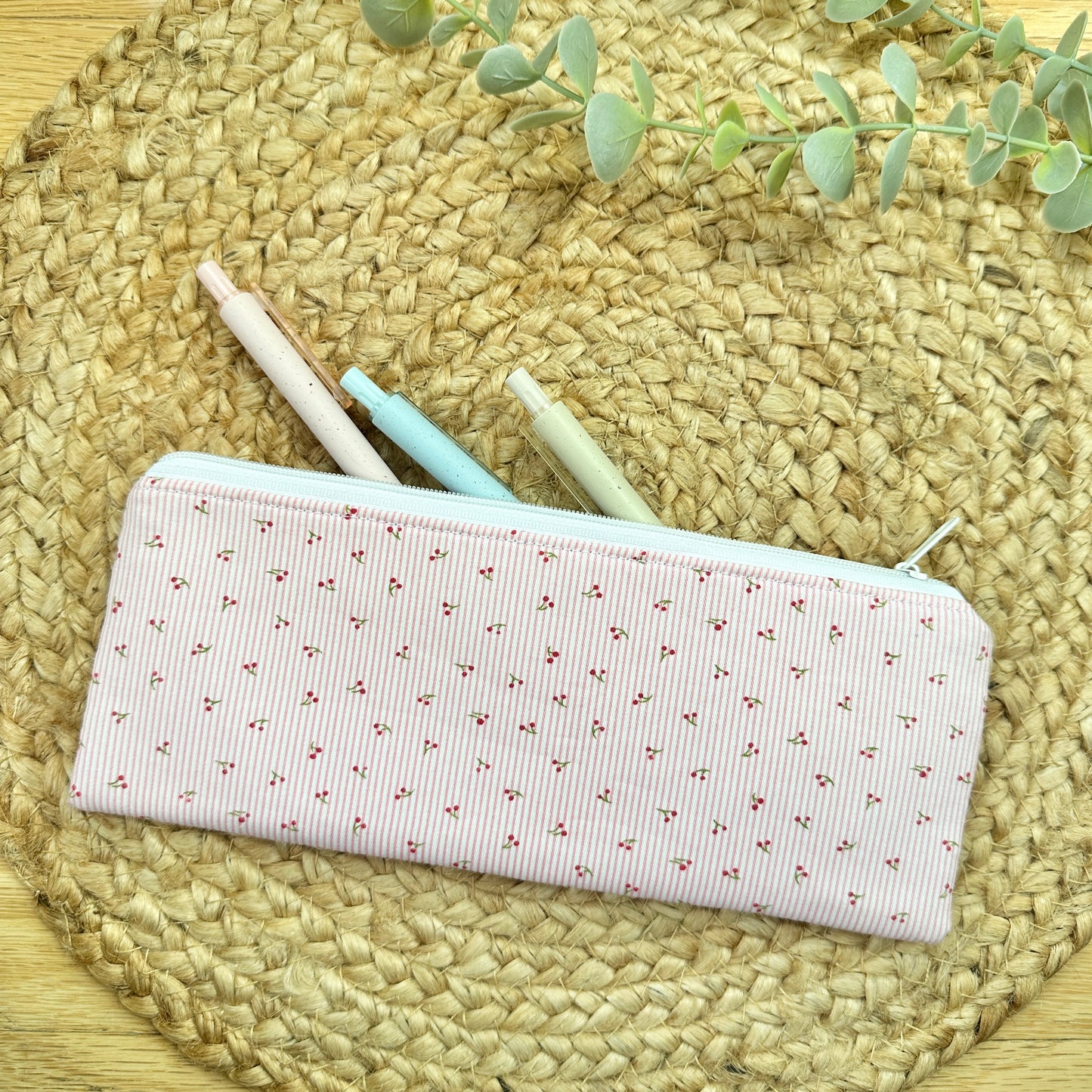 A rectangular fabric pencil case with a light pink and white striped pattern of tiny red hearts, shown on a woven straw mat with three pastel pens peeking out.