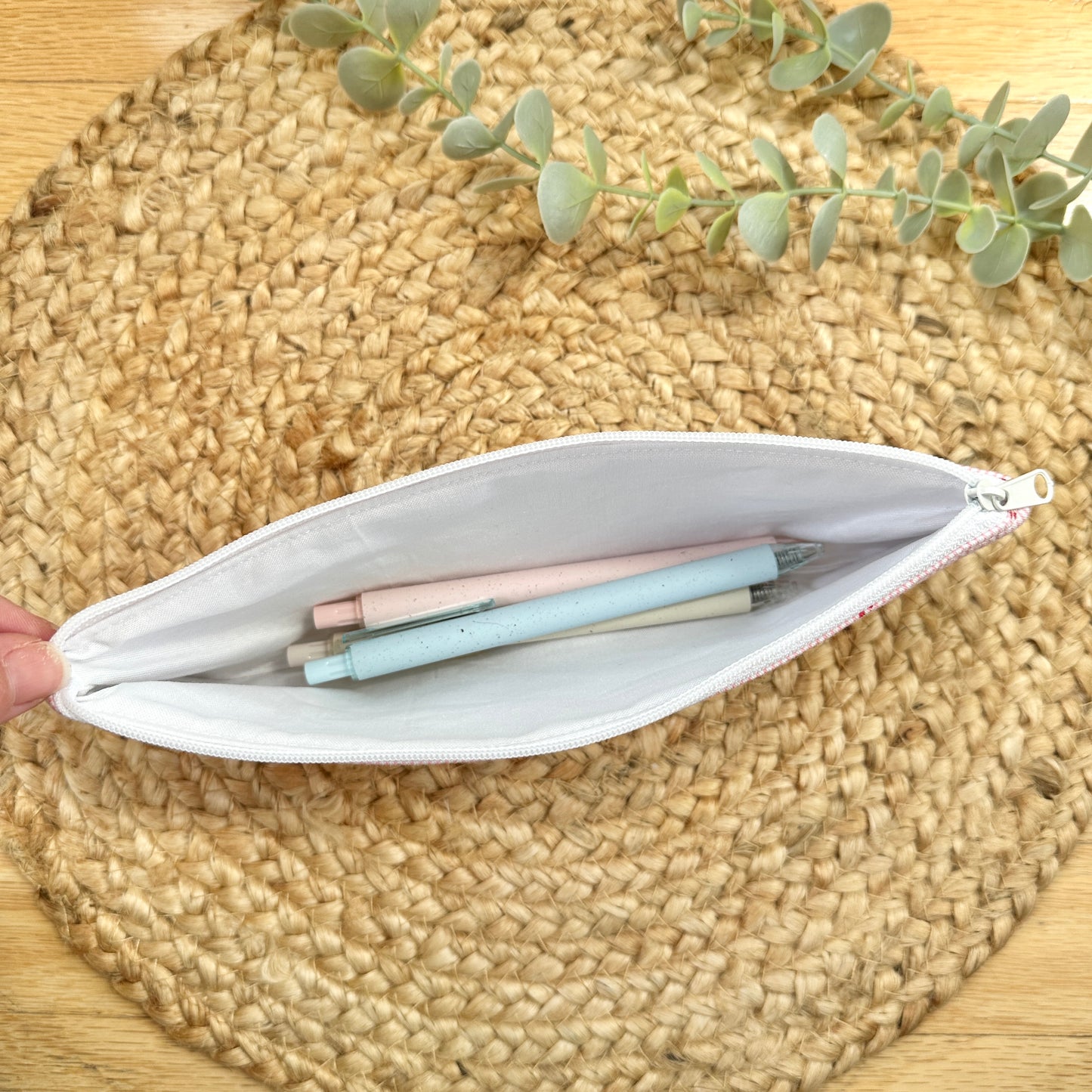 A zipper pouch with a white interior, shown open on a woven straw mat with pastel pens inside.