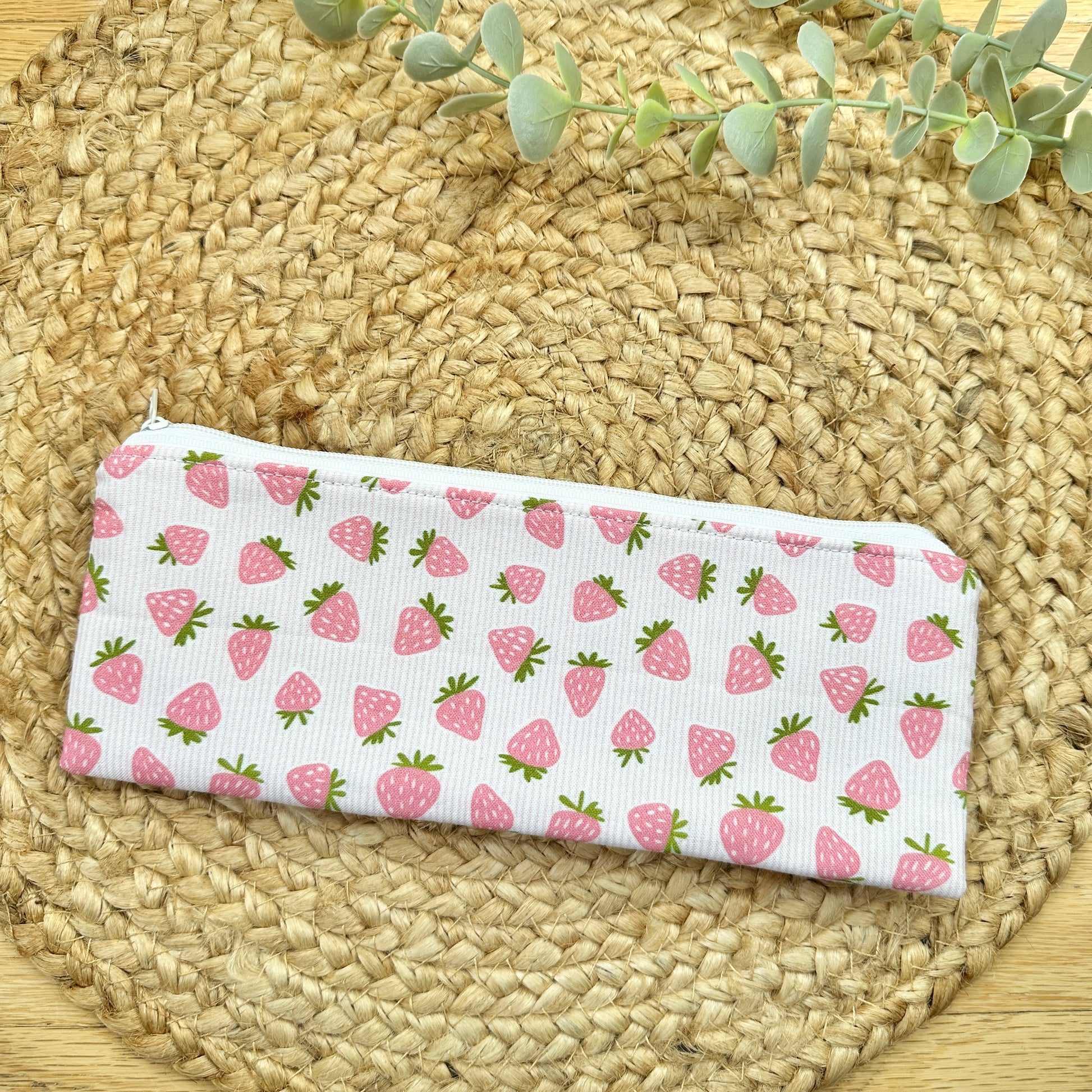 A zipper pencil pouch with a pink strawberry pattern, placed on a round woven straw mat with green leaves at the top.