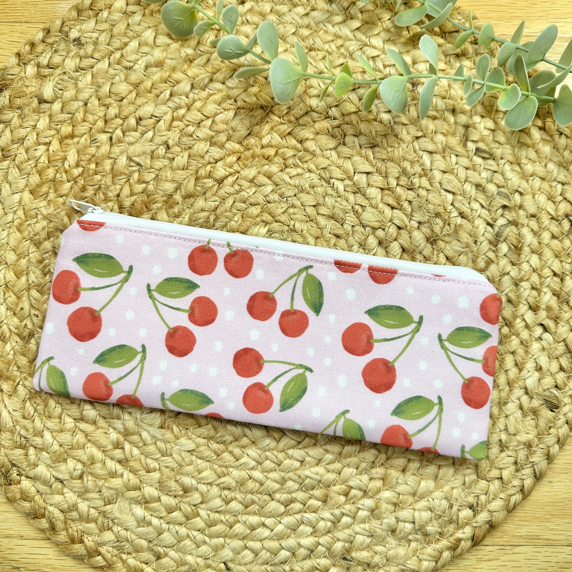 A zippered pencil case with a red cherry and pink pattern, resting on a round woven straw placemat with eucalyptus sprigs in the background.