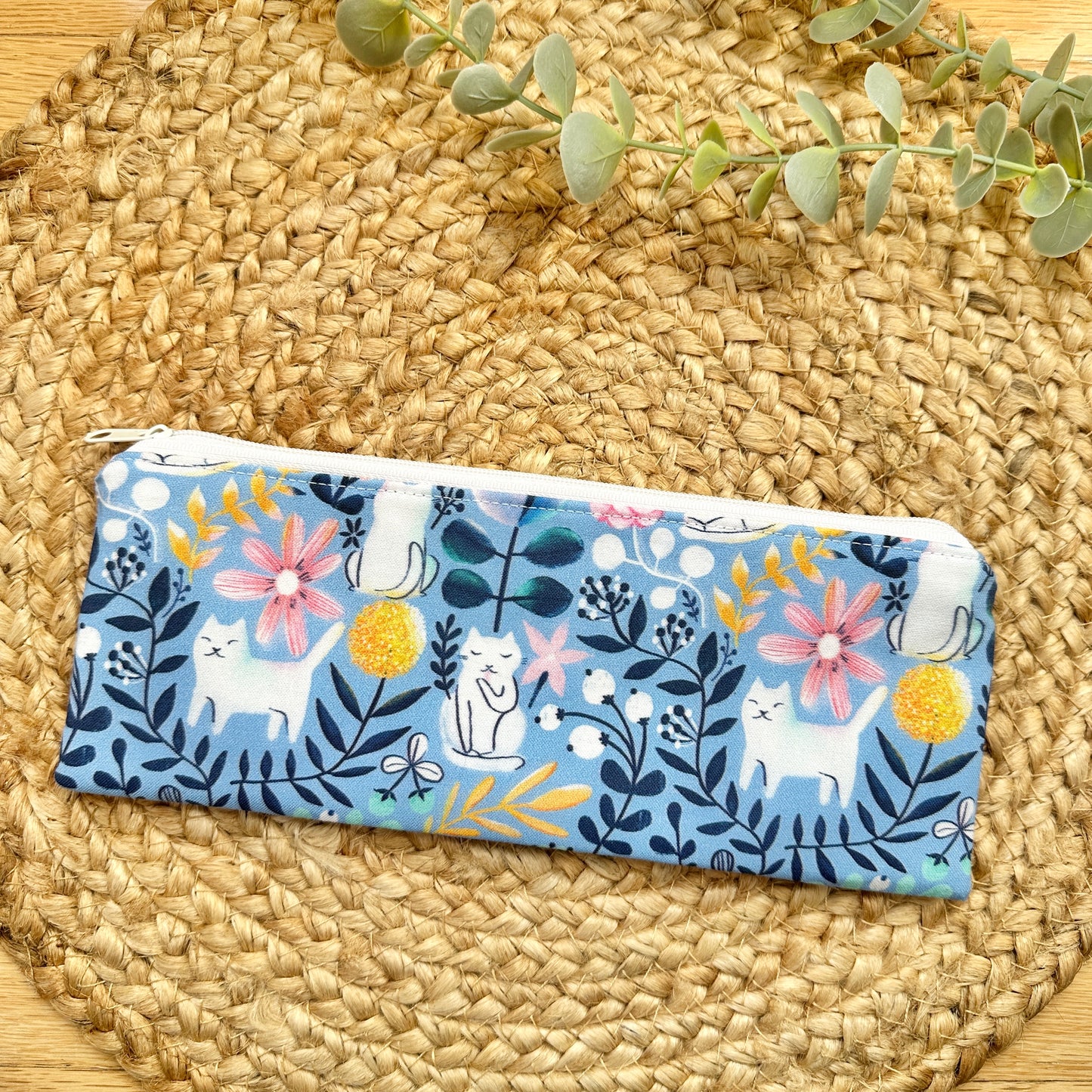 A rectangular zippered pouch with a blue background and a whimsical print of white cats, pink flowers, yellow pom-poms, and dark blue leaves.